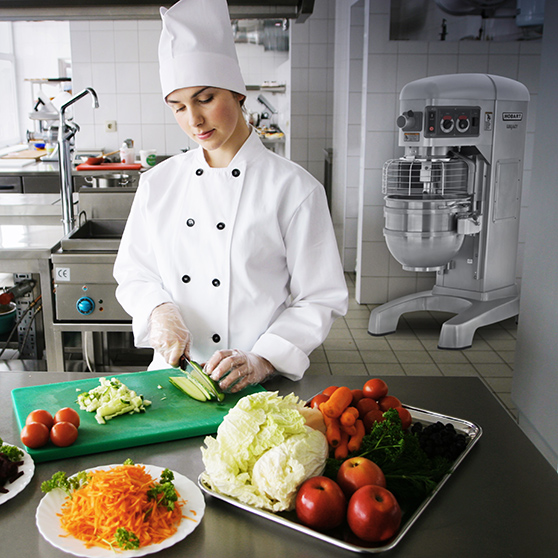 Female chef in a modern commercial kitchen chopping fresh vegetables on a green cutting board, surrounded by tomatoes, carrots, cabbage, and apples, with a HOBART planetary mixer visible in the background next to stainless steel work surfaces.